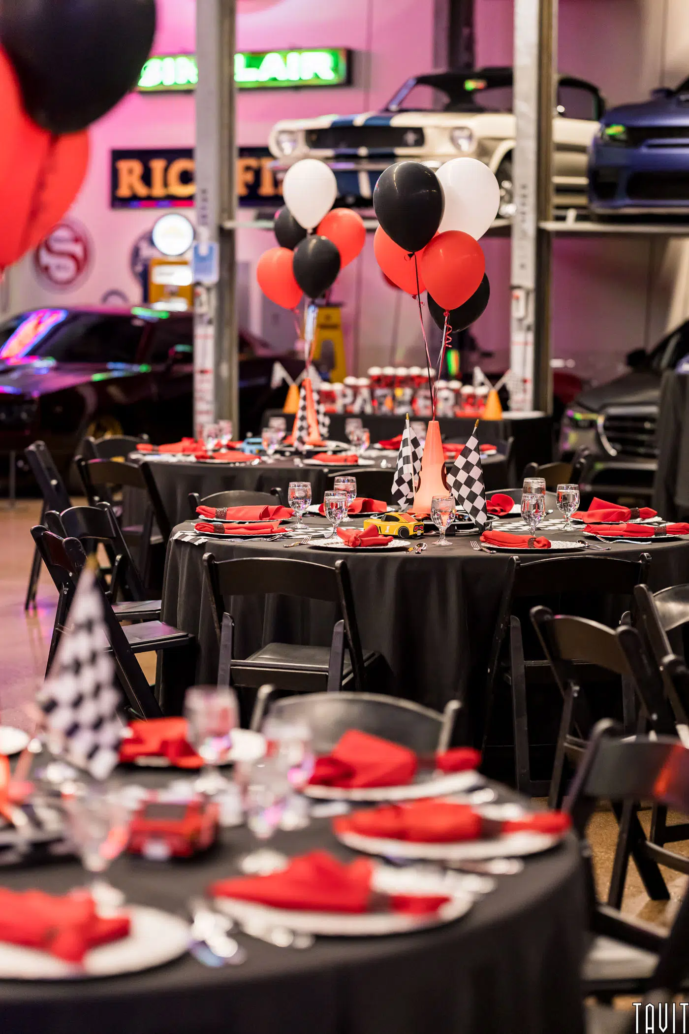 Round tables with black tablecloths, red napkins, and checkered flags are set for a corporate event. Black and red balloons serve as centerpieces, while classic cars and neon signs create a vibrant backdrop.