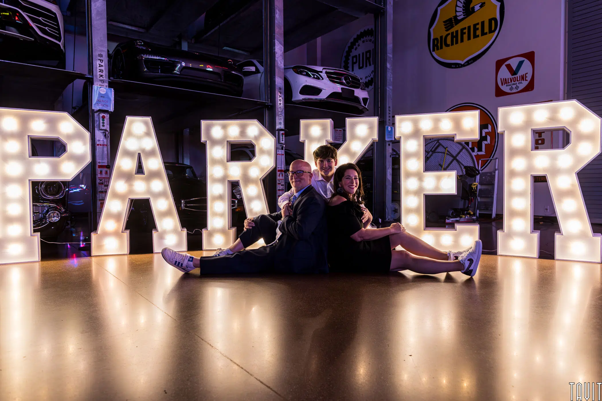 Three people sit on the floor, back-to-back, in front of large illuminated letters spelling "PARKER" inside a garage with vintage cars and retro signs—creating a unique atmosphere for a corporate event or seminar.