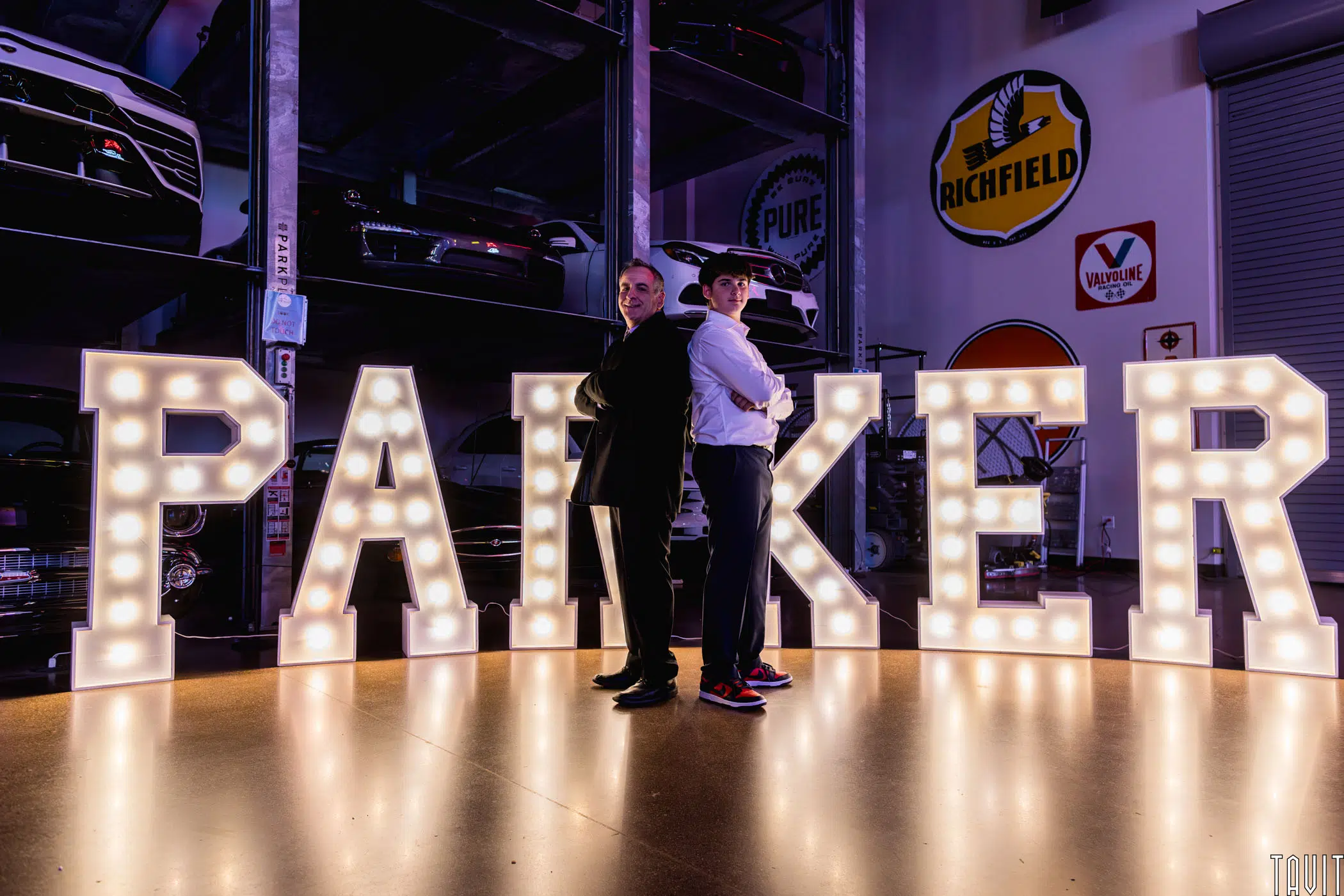 Two people in formal attire stand back to back in front of large illuminated letters spelling "PARKER," at a corporate event, with classic cars and vintage signs visible in the background.