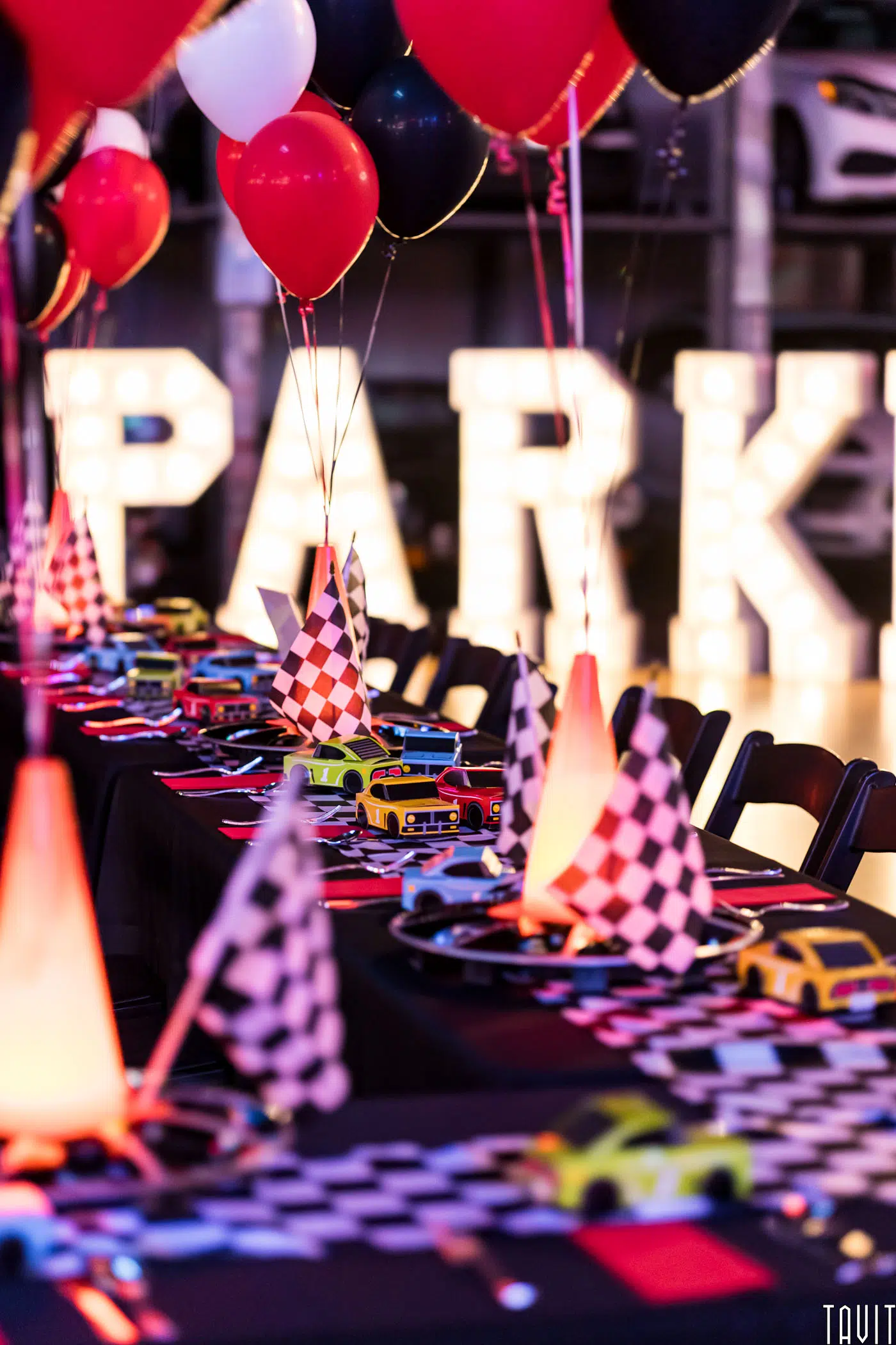 A long table decorated with checkered racing flags, toy cars, and red and black balloons sets a festive scene at the corporate event. In the background, large illuminated letters spell out "PARK," adding to the colorful atmosphere.