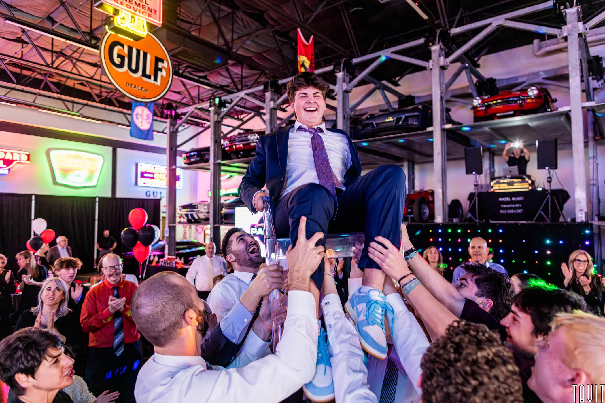 A young man in a suit and tie is lifted on a chair by a group of people at a lively corporate event in a decorated space with vintage cars and neon signs. The crowd looks joyful and festive.