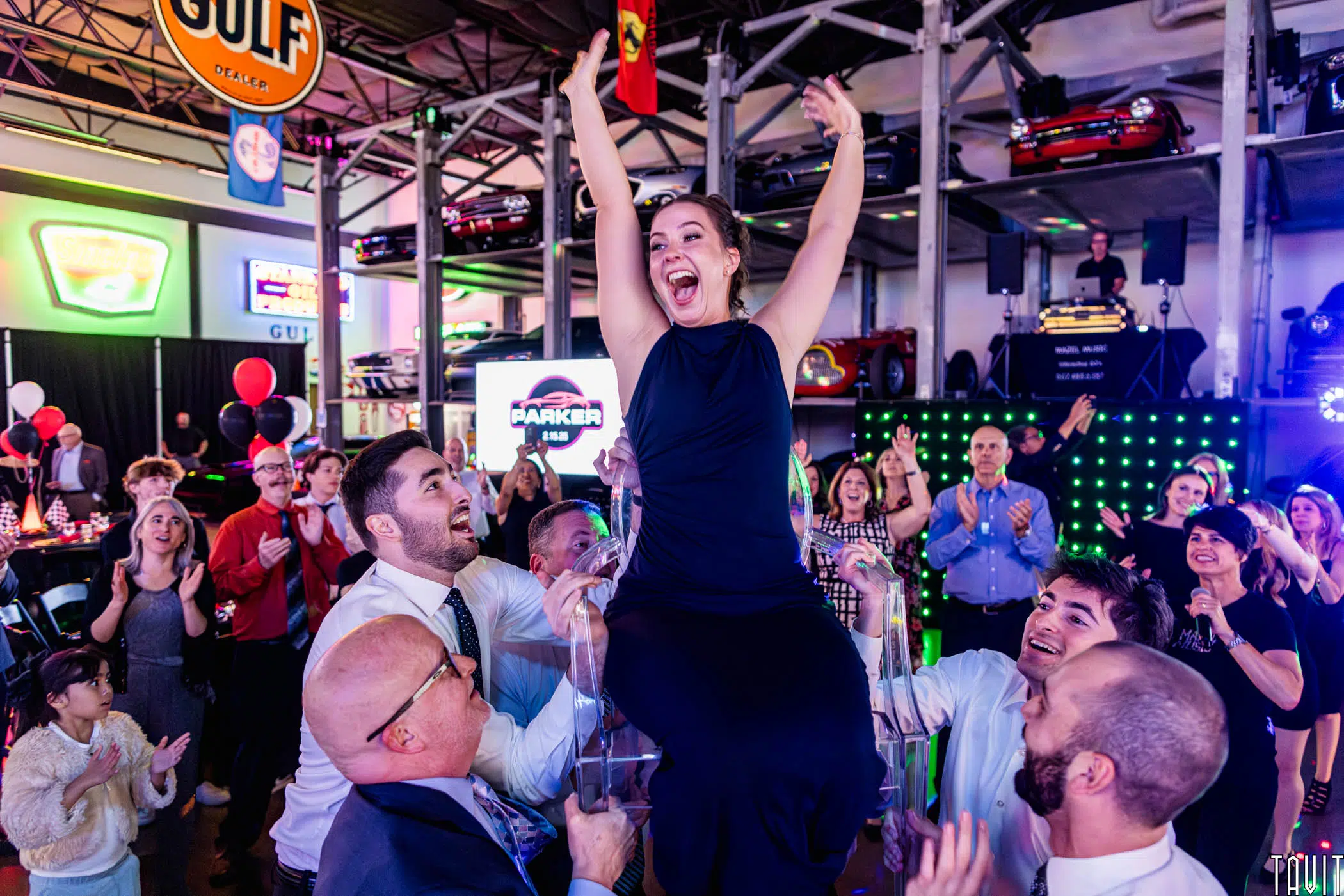 A woman in a black dress is lifted on a chair by four men at a lively corporate event, surrounded by cheering guests in a car-themed venue with colorful lights and vintage car decor.