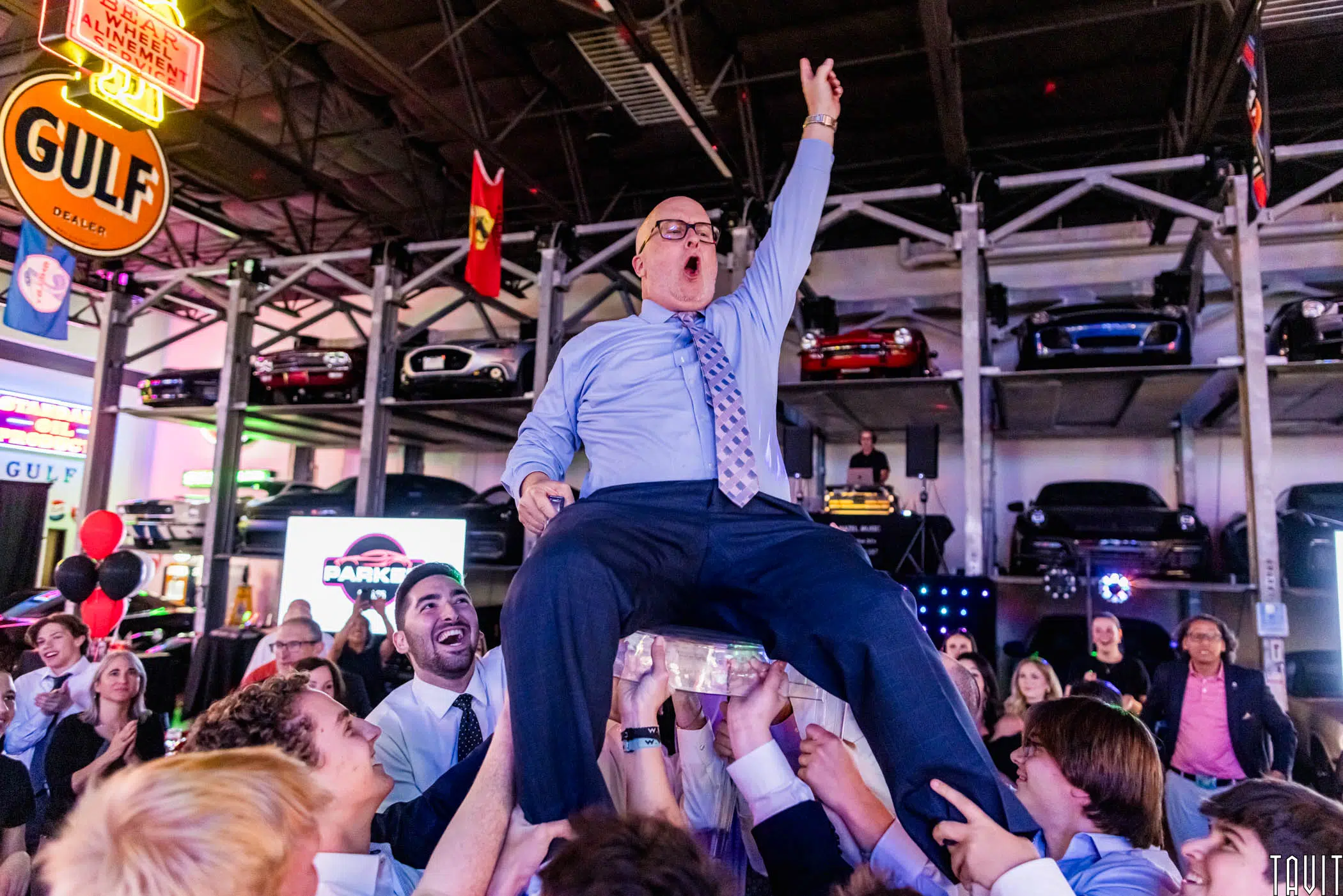 A jubilant man in a suit and tie is lifted on a chair by a cheering crowd at a lively corporate event, with classic cars and neon signs in the background.