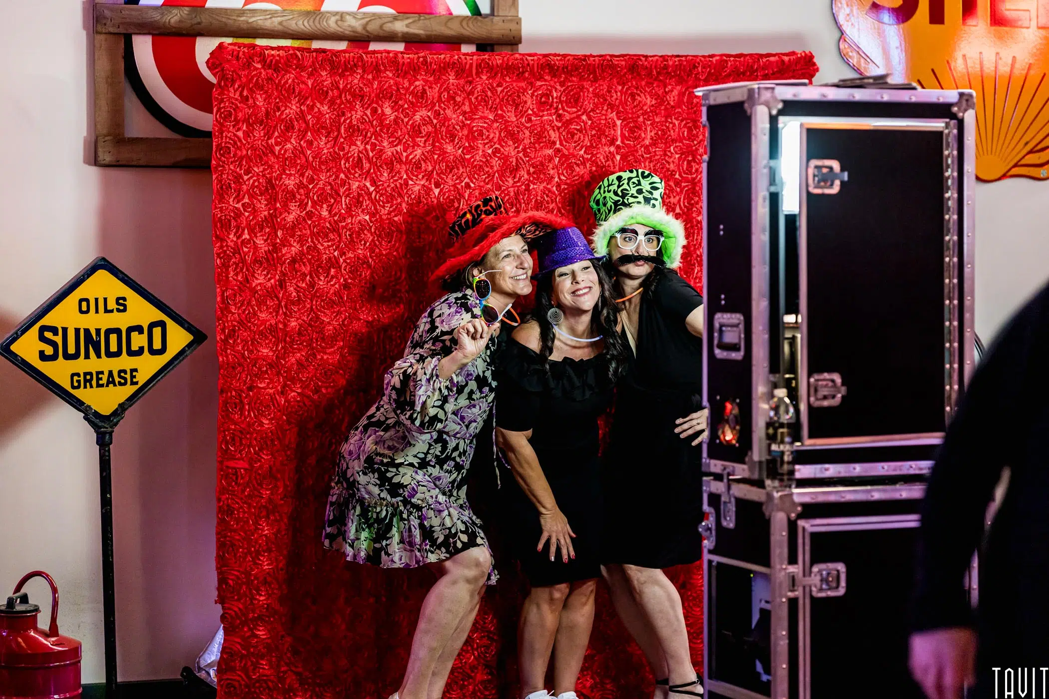 Three women in fun hats and fake mustaches pose together near a vintage Sunoco sign and photo booth setup, bringing playful party vibes to this corporate event with a red flower-patterned backdrop.
