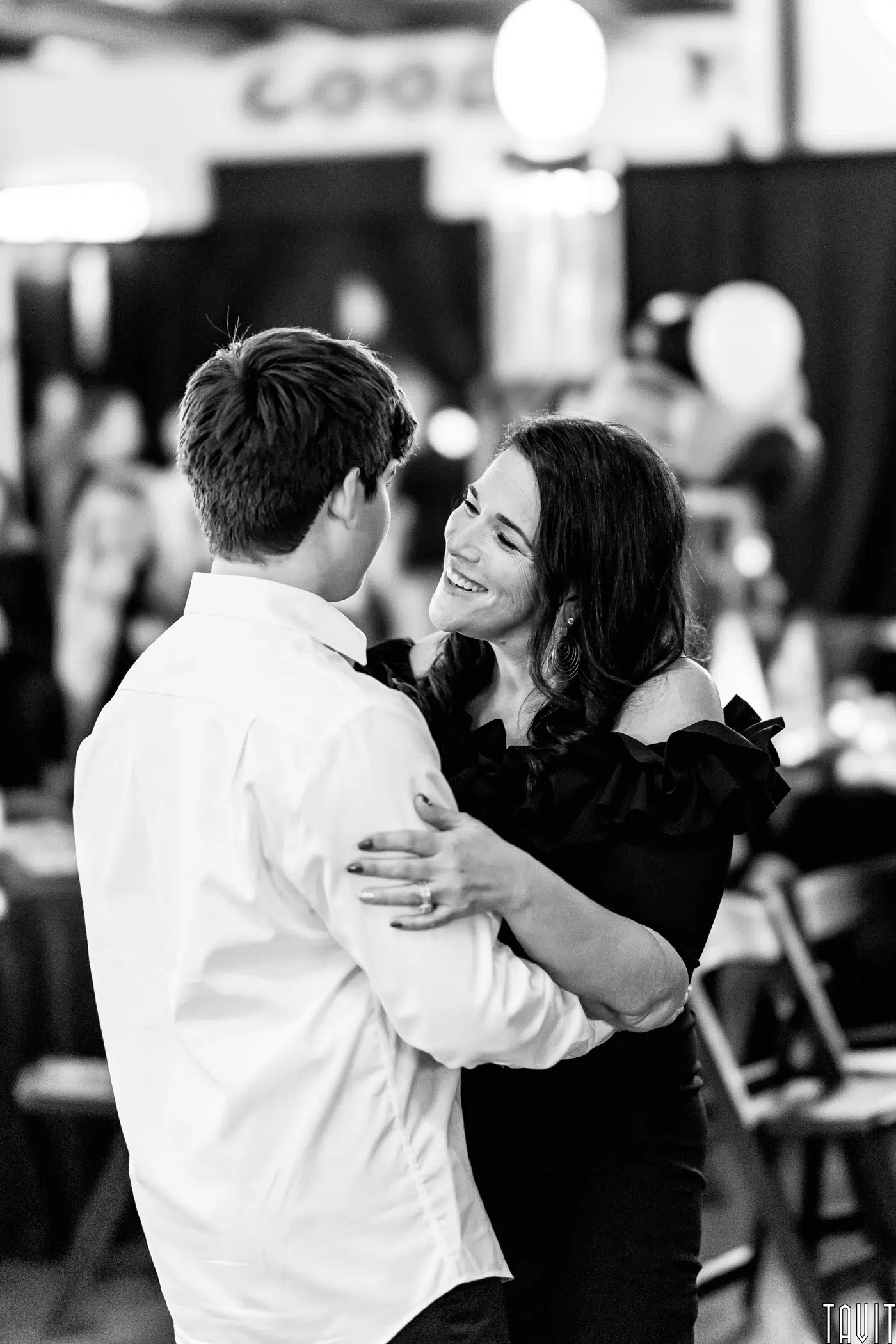 A woman in a black off-the-shoulder dress smiles warmly while dancing with a young man in a white shirt at a lively corporate event, softly lit with blurred guests and elegant decorations in the background.