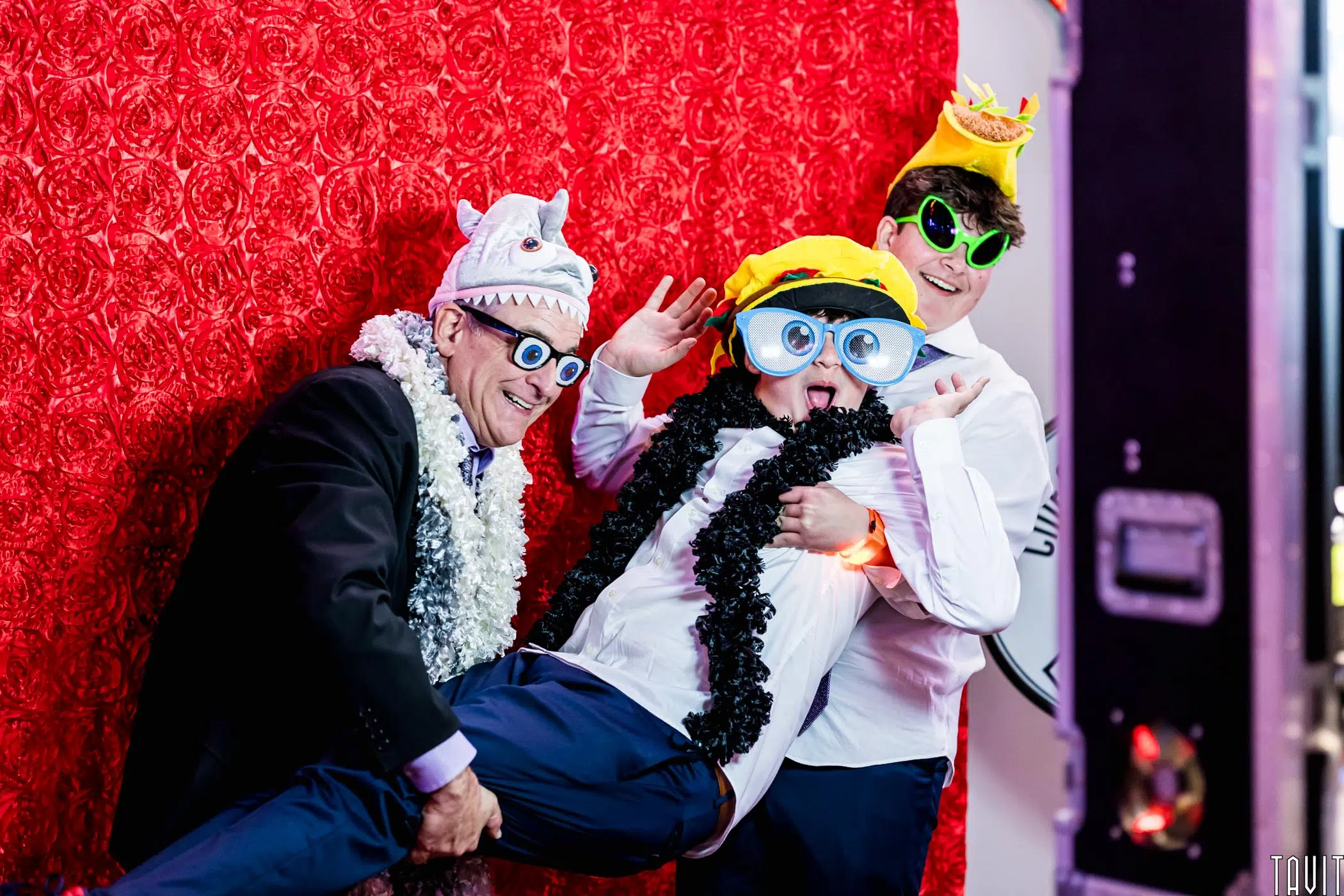 Three people in playful costumes and oversized glasses pose in front of a red floral backdrop at a corporate event; one man holds up a laughing boy while another person smiles, all wearing hats and feather boas.