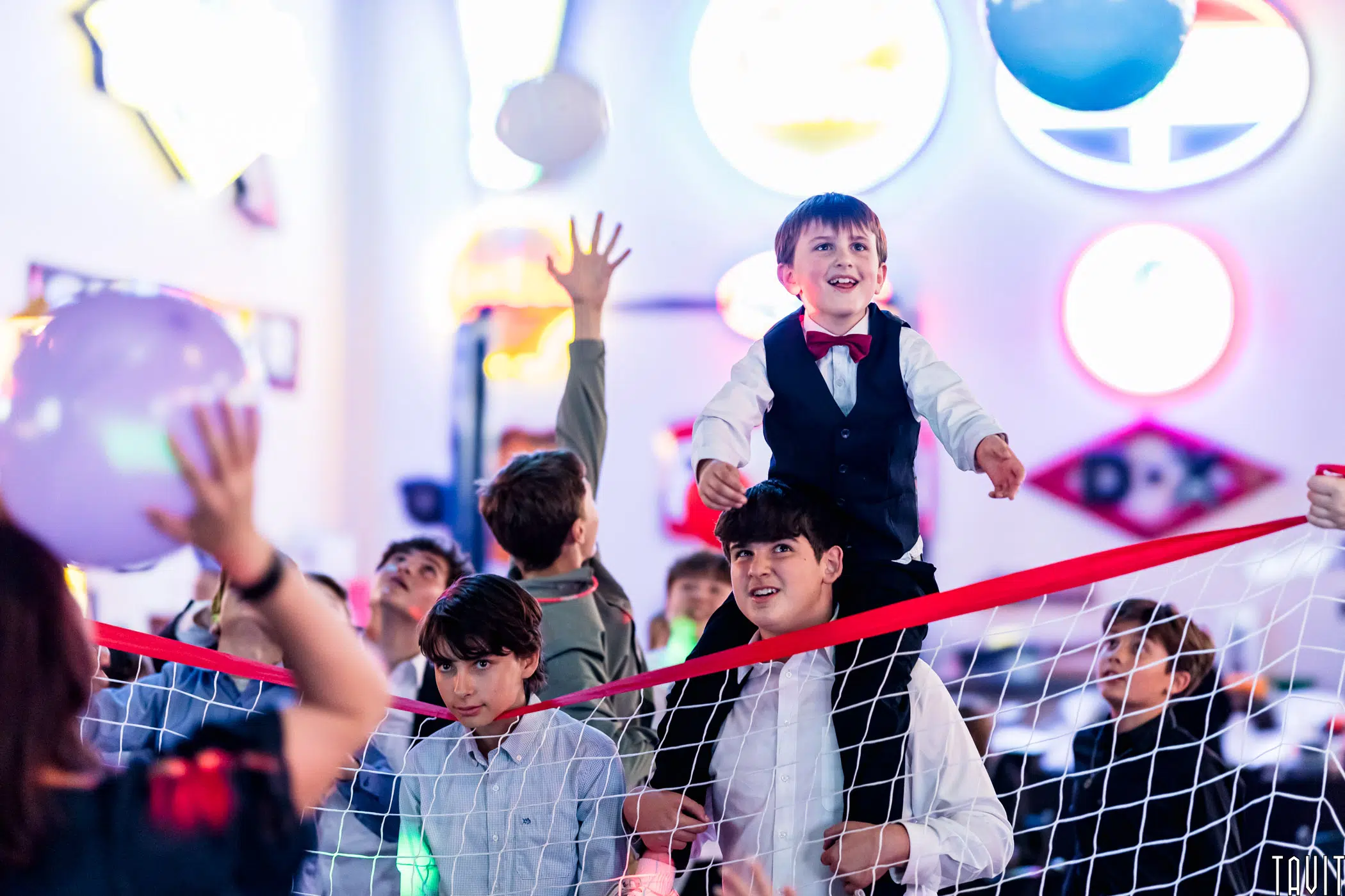 Children play with balloons at a party. One boy in a vest and red bow tie sits on another’s shoulders near a net, as colorful, blurred lights create an atmosphere more lively than any corporate event or seminar.
