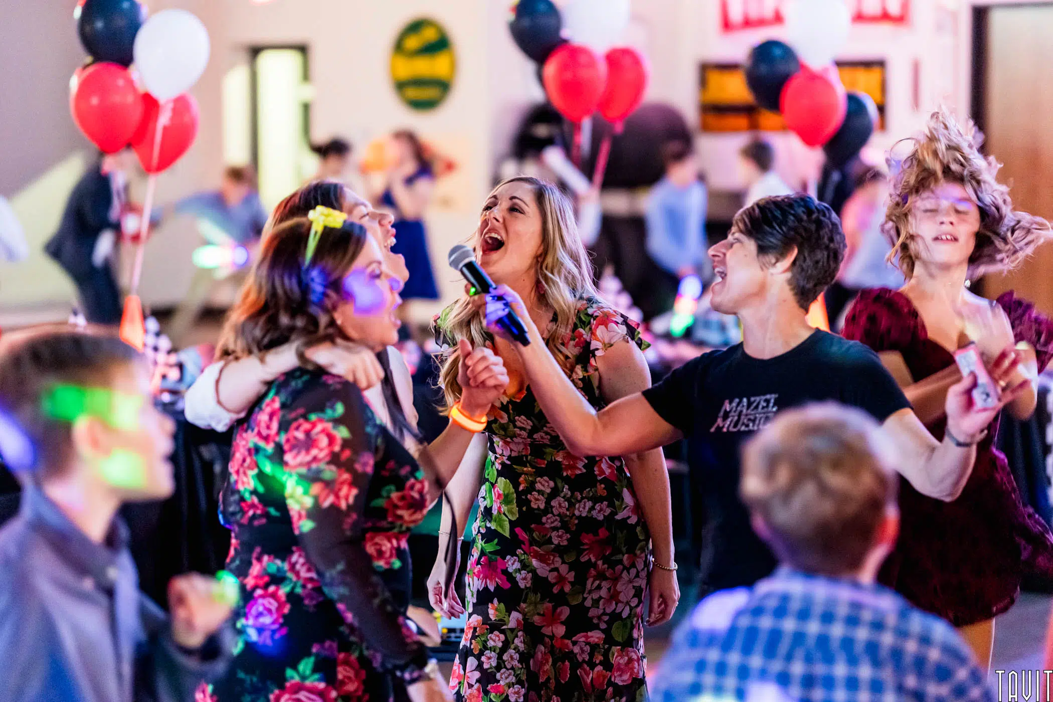 A group of people sing enthusiastically into a microphone at a lively corporate event, surrounded by balloons and colorful lights. The atmosphere is joyful and energetic, with others dancing and celebrating in the background.