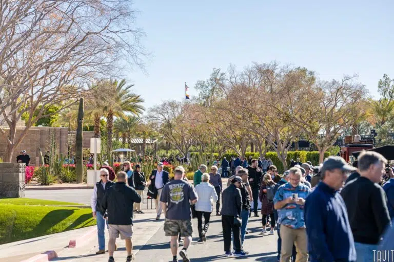 A crowd of people walks along a sunny, tree-lined street in a park-like setting, many wearing jackets and hats—perhaps attending an outdoor expo. Palm trees and leafless trees line the path, with a flag flying under a clear blue sky.