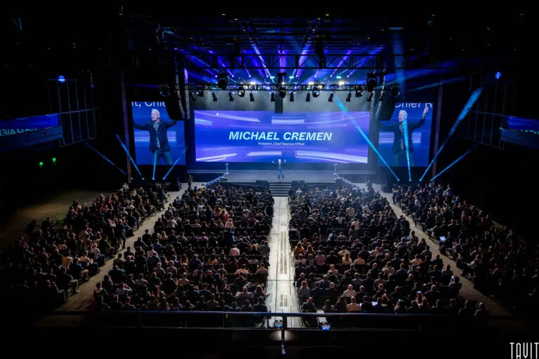 A large audience sits in a dark auditorium at a corporate event, facing a brightly lit stage where a man speaks in front of a screen displaying the name "Michael Cremen." Blue lighting and projections illuminate the scene.