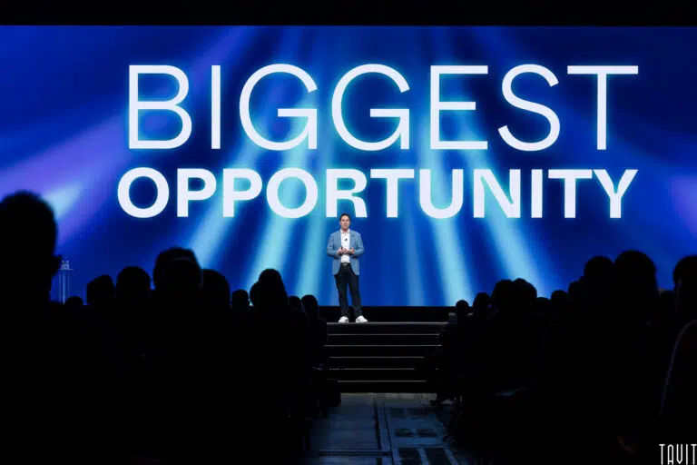 A speaker stands on stage at a corporate event, in front of a large screen displaying the words "BIGGEST OPPORTUNITY," while an audience watches in a darkened auditorium.