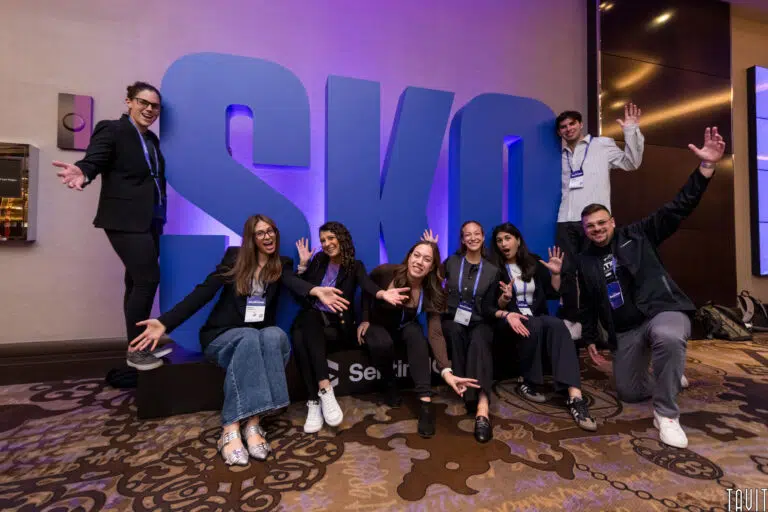 A group of eight people pose enthusiastically in front of large blue "SKO" letters at a lively corporate event, smiling and gesturing with open arms. The professional indoor setting resembles a vibrant expo atmosphere.