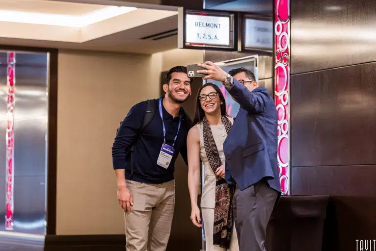 Three people stand together in a hallway, smiling as one takes a selfie at a corporate event. Name badges are visible, suggesting they're attending a seminar or expo. The background features a sign labeled "Belmont 1, 2, 5, 6.