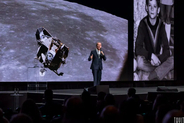A man in a suit speaks on stage with a microphone at a corporate event. Behind him, a large screen displays a lunar module above the moon and a black-and-white photo of a boy, with audience silhouettes visible in the foreground.
