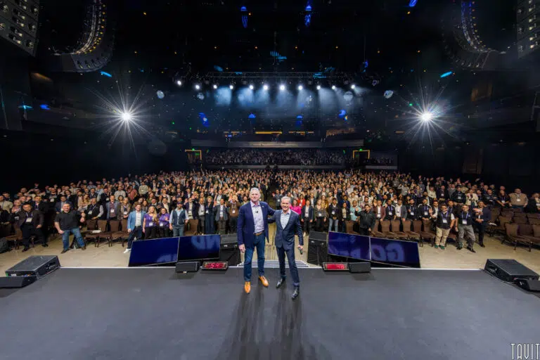 Two men stand on a stage at a corporate event, facing a large, seated audience in a dimly lit auditorium. Bright stage lights shine overhead as the attentive crowd focuses on the speakers.