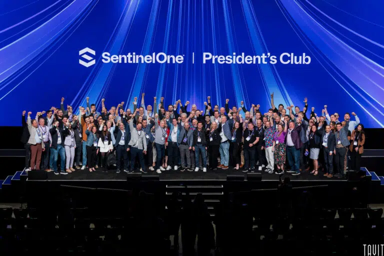 A large group of people pose together on stage under a blue backdrop with the "SentinelOne | President’s Club" logo at this corporate event, many raising their fists and smiling for the camera.