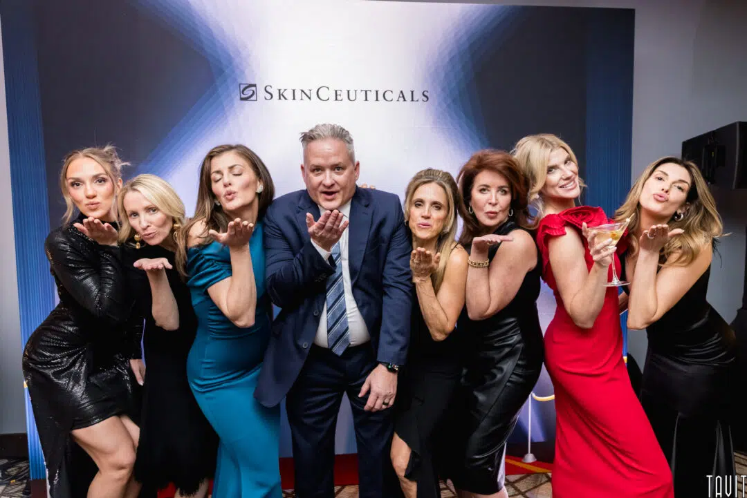 A group of seven people, six women and one man, dressed in formal attire, stand in front of a SkinCeuticals backdrop at a corporate event. Most are blowing kisses toward the camera and smiling, creating a fun and lively atmosphere.