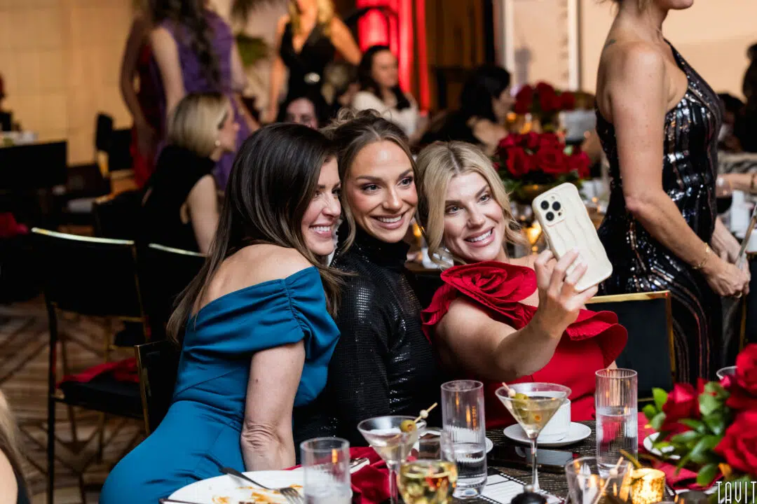 Three women dressed formally sit at a dinner table during a corporate event, smiling and posing for a selfie. The table is set with drinks and glasses, while other guests mingle in the background.
