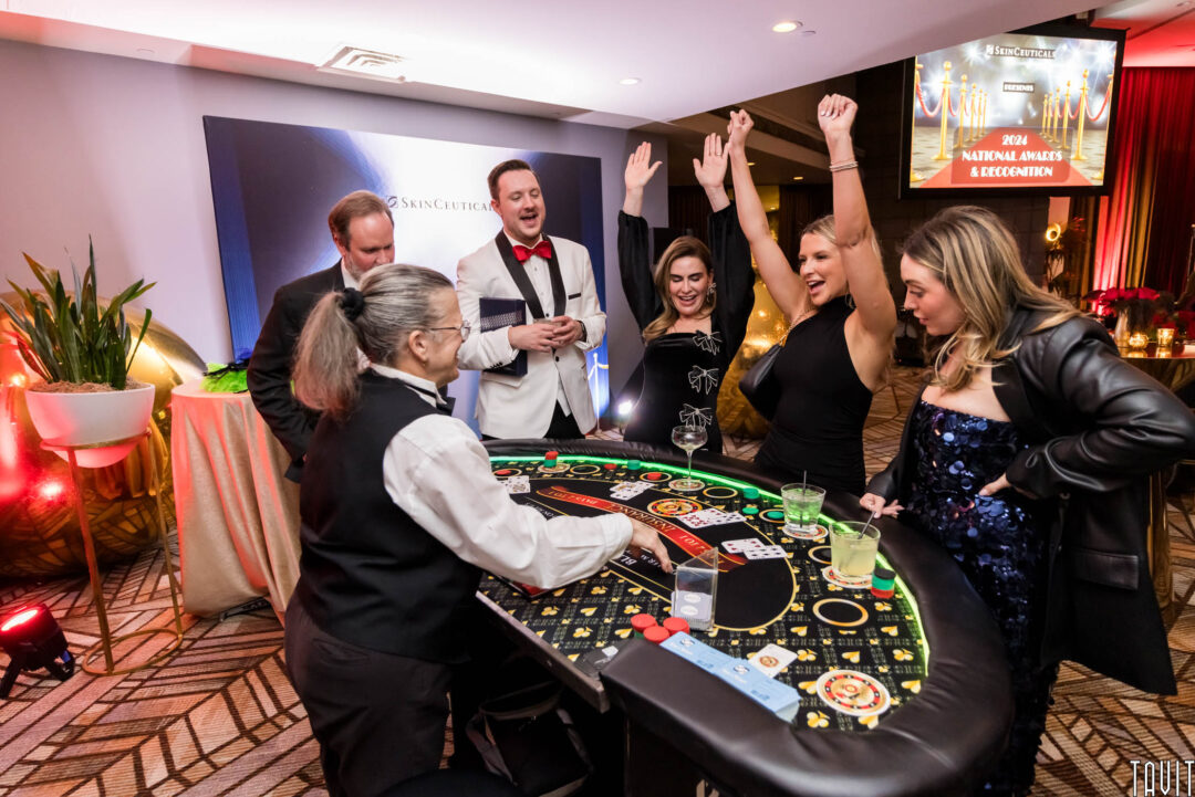 Six people gather around a casino table at a corporate event, dressed in formal attire. One woman cheers with arms raised, celebrating a win. The dealer smiles as colorful lights and a screen displaying "2024 National Awards & Recognition" shine in the background.