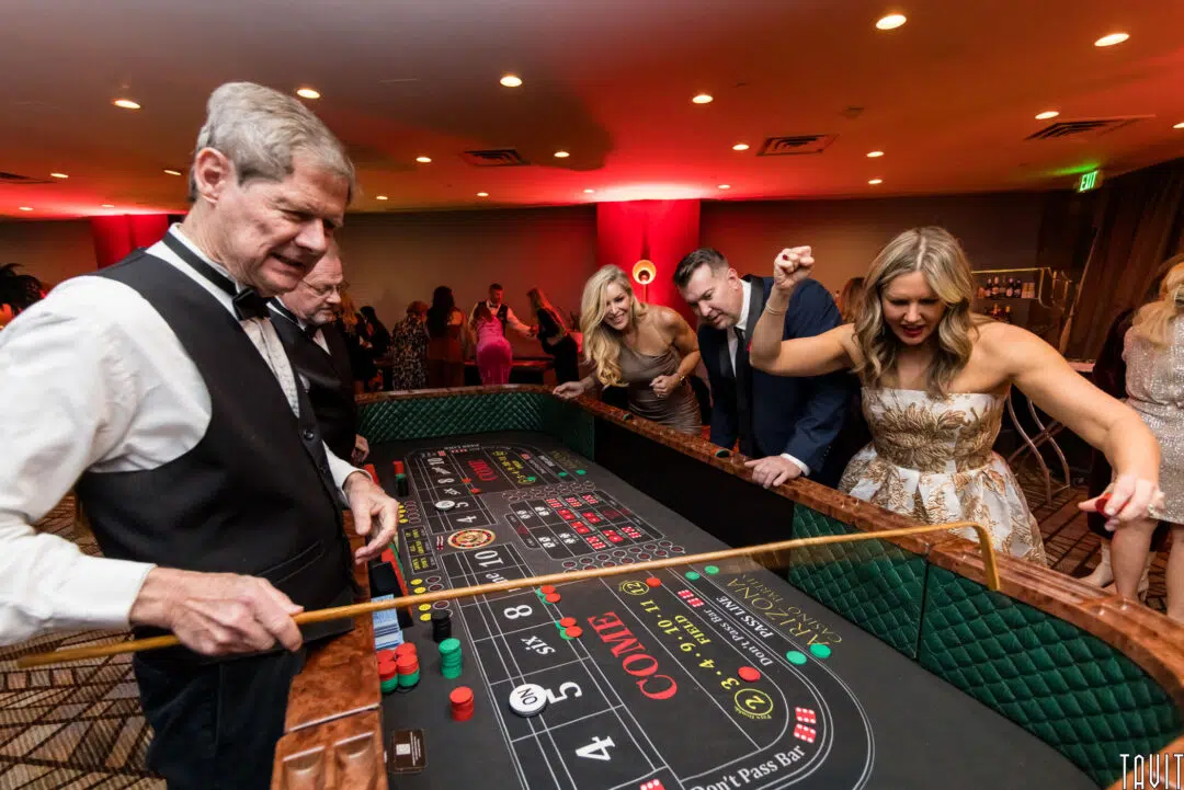 A group of people dressed in formal attire play craps at a corporate event casino table. A woman on the right is excited, raising her arm, while a dealer in a black vest supervises. Other guests watch and smile in the background.