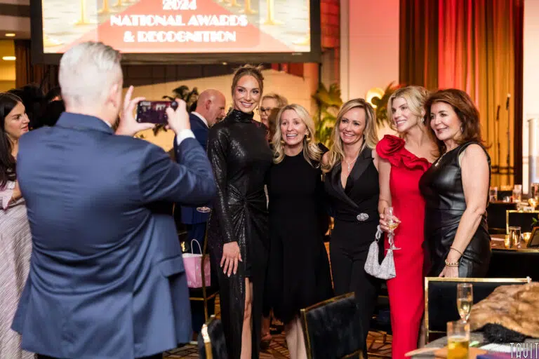 A group of five women pose and smile as a man takes their photo with his phone at a formal corporate event. A sign in the background reads "2024 National Awards & Recognition.