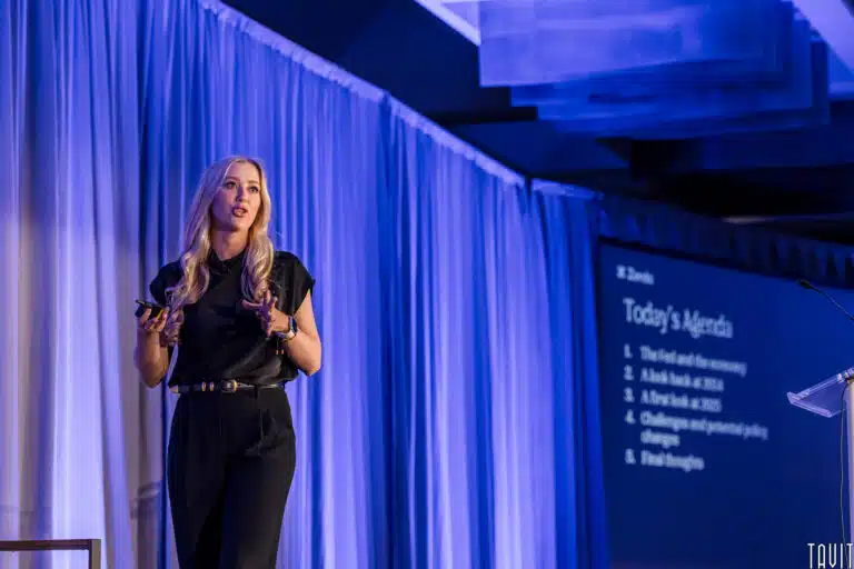 A woman with long blond hair stands on stage giving a presentation. She is wearing a black outfit and holding a remote. A presentation slide behind them displays text, including "Today's Agenda" and a list of topics.