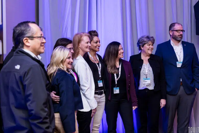 A group of professionally dressed people pose together and smile for a photo at an indoor corporate event, with a blue-lit curtain in the background.