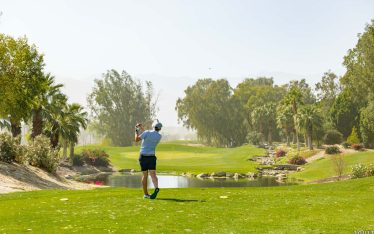 A golfer wearing shorts and a hat swings a club on a lush, green golf course bordered by palm trees, with a small pond and mountains visible in the background under a hazy sky.