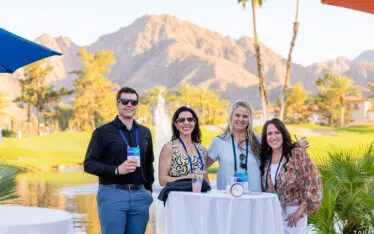 Four people stand and smile at a high-top table outdoors by a pond, with palm trees and mountains in the background on a sunny day. They wear conference badges and sunglasses, and drinks are on the table.