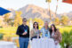 Four people stand and smile at a high-top table outdoors by a pond, with palm trees and mountains in the background on a sunny day. They wear conference badges and sunglasses, and drinks are on the table.