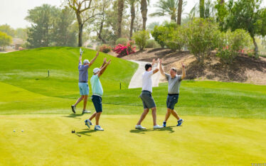 Four men on a sunny golf course celebrate with high fives and raised arms, surrounded by lush green grass, palm trees, and colorful flowering bushes.