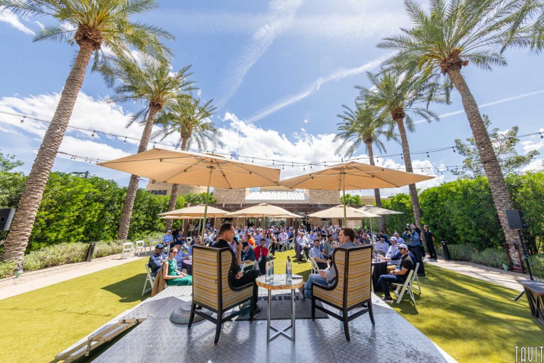 An outdoor panel discussion is taking place under large umbrellas, surrounded by palm trees and greenery. Captured by a professional Event Photographer in Phoenix, audience members sit on chairs, enjoying the sunny weather.