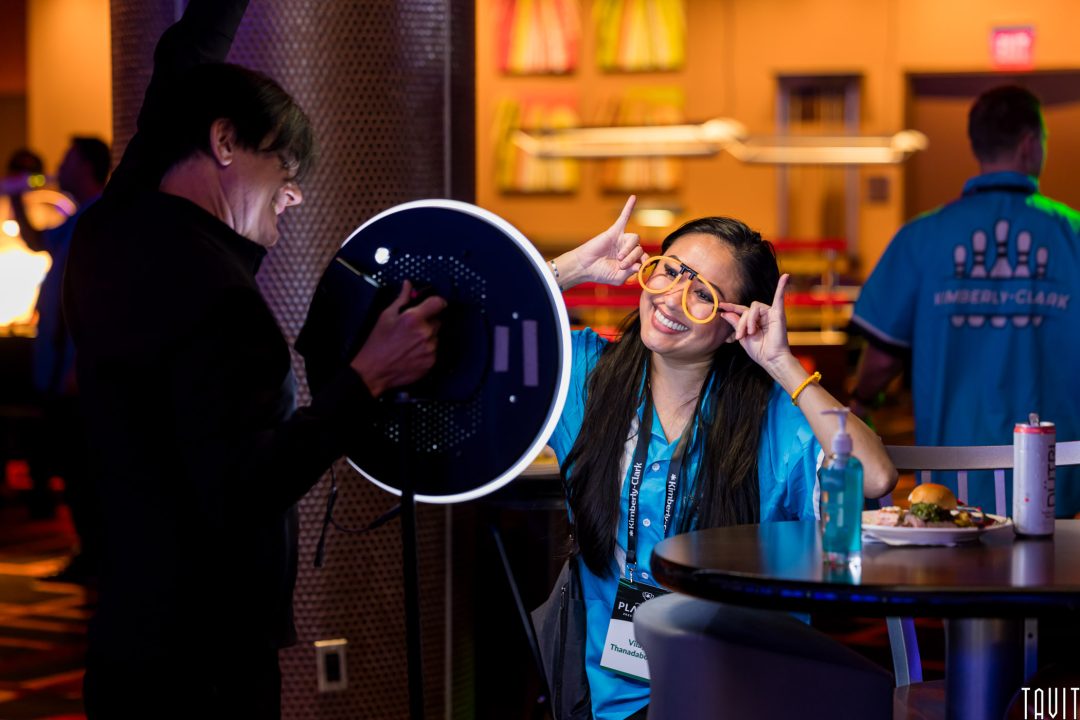 A woman wearing orange glasses and a blue shirt poses playfully with her fingers by her head while being photographed by a Corporate Event Photographer Phoenix with a ring light at an indoor event; food, drinks, and hand sanitizer are on a nearby table.