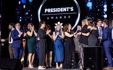 A group of people dressed formally stand on stage clapping and smiling in front of a large sign that reads "President's Awards," with colorful lights in the background.