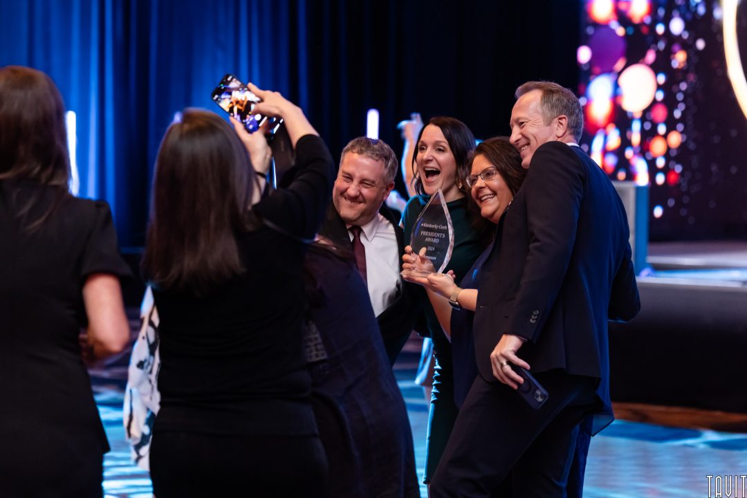 A group of people in formal attire pose together and smile for a photo at an awards event, holding a trophy. Captured by a skilled Corporate Event Photographer, the scene features blue lighting and a screen with colorful lights in the background.