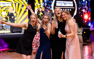 Four women in dresses pose and smile for a photo at an indoor event, holding drinks, with a bright, colorful stage and a "Customer Development Excellence Awards" sign in the background.