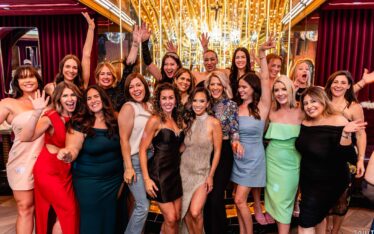 A group of women dressed in colorful, stylish dresses pose together indoors, smiling and waving excitedly at a Scottsdale Holiday Party Photos event in front of a gold-lit backdrop with reflective surfaces and vibrant decor.