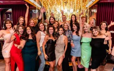 A group of women dressed in colorful, stylish dresses pose together indoors, smiling and waving excitedly at a Scottsdale Holiday Party Photos event in front of a gold-lit backdrop with reflective surfaces and vibrant decor.