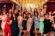 A group of women dressed in colorful, stylish dresses pose together indoors, smiling and waving excitedly at a Scottsdale Holiday Party Photos event in front of a gold-lit backdrop with reflective surfaces and vibrant decor.