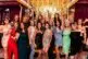 A group of women dressed in colorful, stylish dresses pose together indoors, smiling and waving excitedly at a Scottsdale Holiday Party Photos event in front of a gold-lit backdrop with reflective surfaces and vibrant decor.