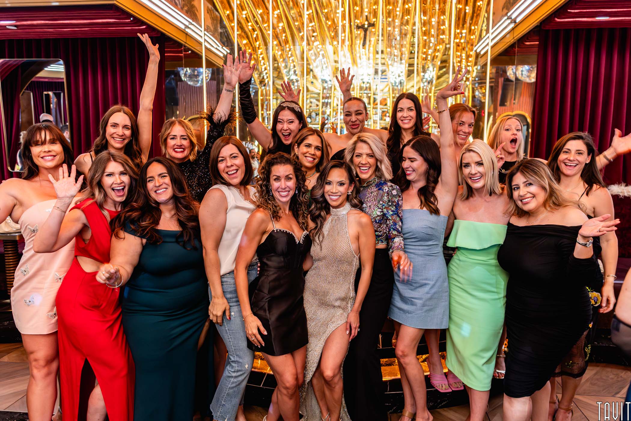 A group of women dressed in colorful, stylish dresses pose together indoors, smiling and waving excitedly at a Scottsdale Holiday Party Photos event in front of a gold-lit backdrop with reflective surfaces and vibrant decor.