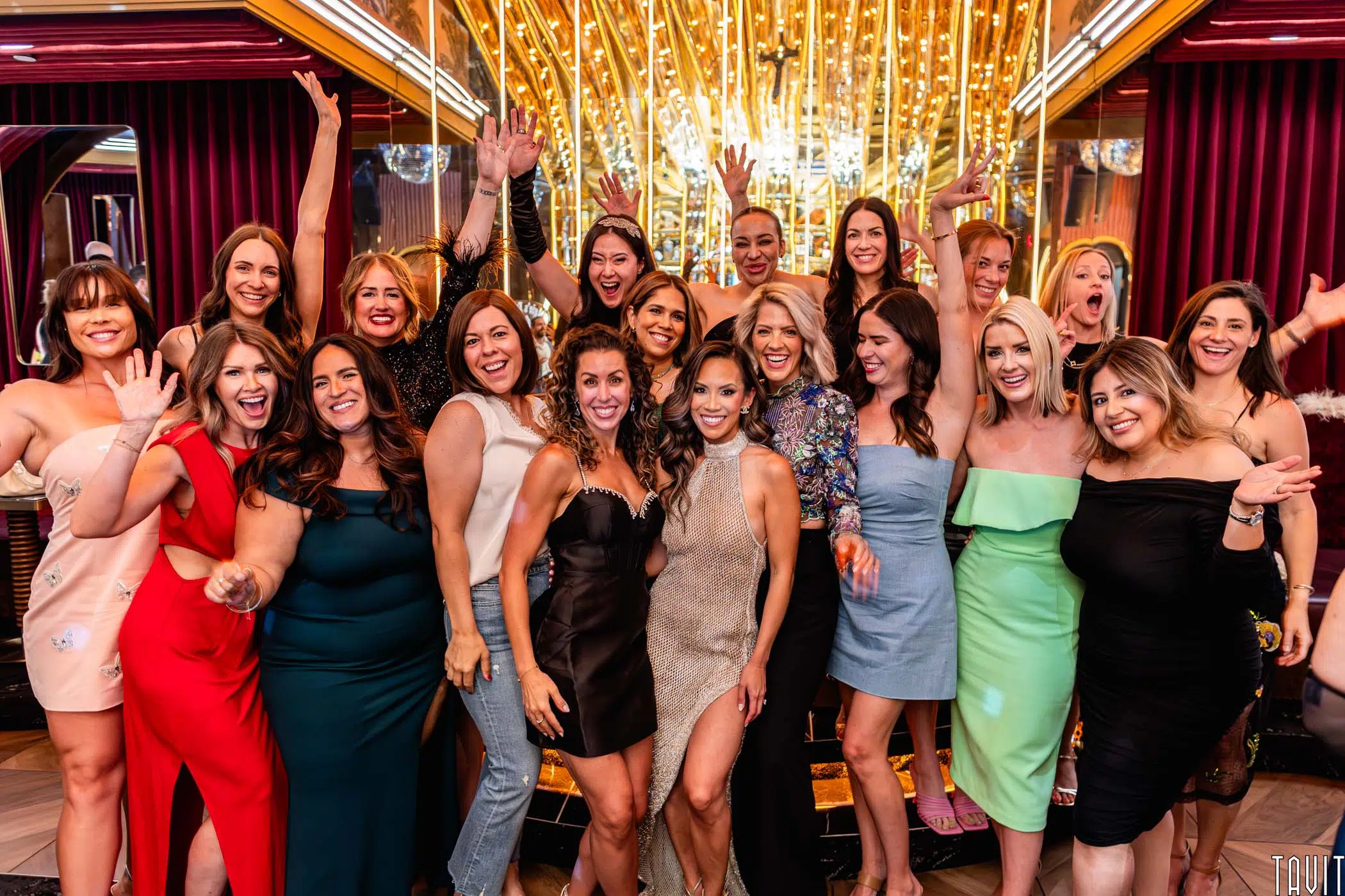 A group of women dressed in colorful, stylish dresses pose together indoors, smiling and waving excitedly at a Scottsdale Holiday Party Photos event in front of a gold-lit backdrop with reflective surfaces and vibrant decor.