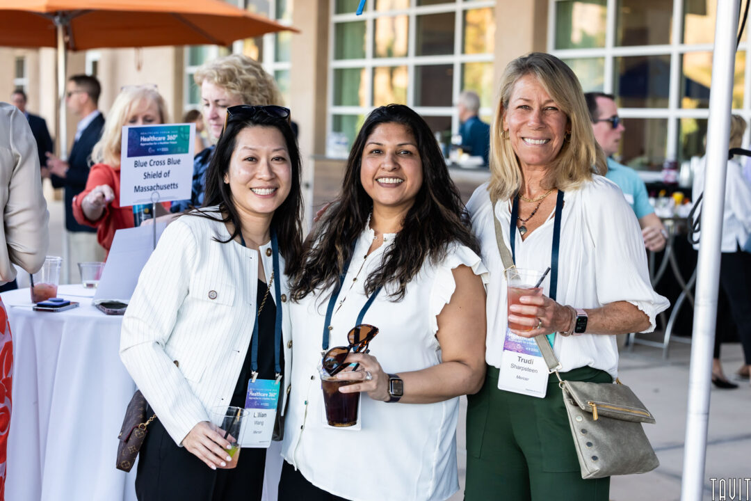 Three women with conference badges smile together at an outdoor networking event. Captured by a Corporate Event Photographer Phoenix, they each hold drinks while business professionals mingle in the background near tables.