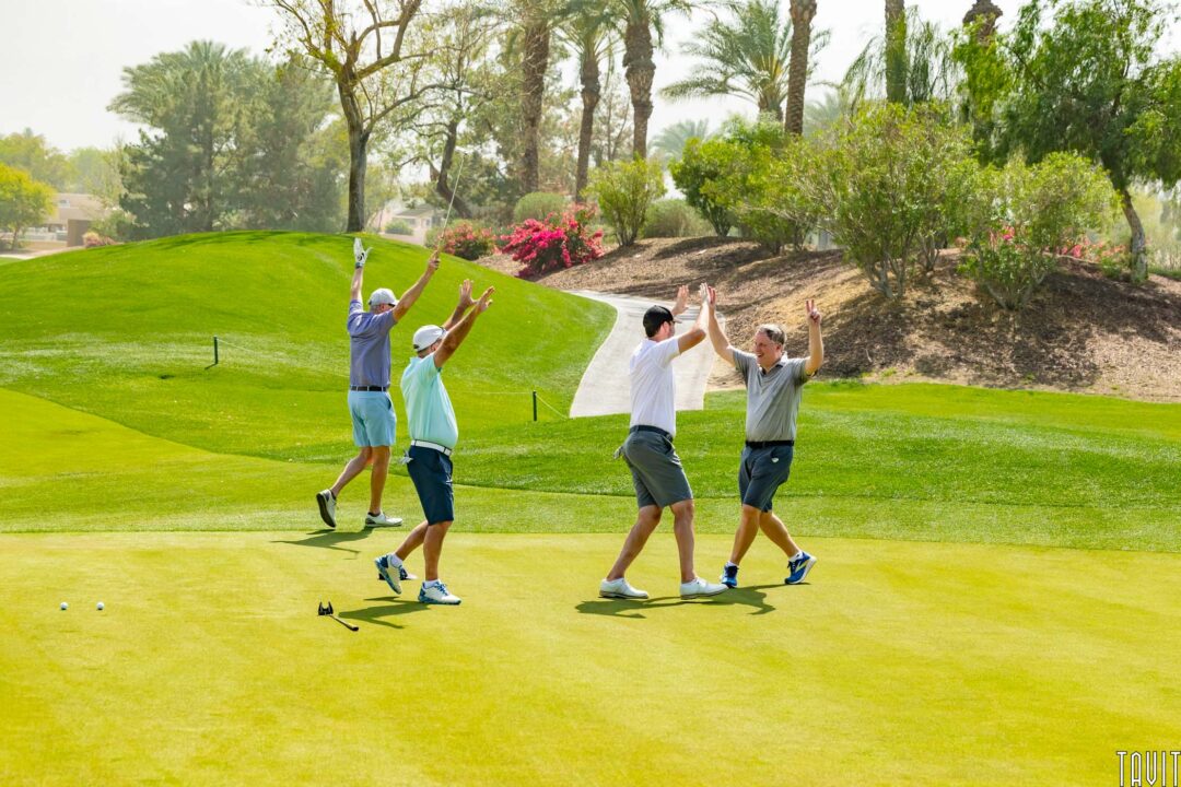 Four men on a sunny golf course celebrate with raised arms and high-fives, surrounded by palm trees and colorful flowers. Event Photography captures the excitement as golf clubs and balls rest nearby on the lush green grass.