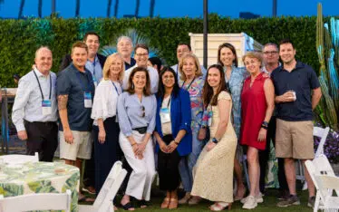 A group of sixteen adults smile and pose together outdoors at an event during the evening, surrounded by plants and palm trees, with tables and white chairs in the foreground—capturing the moment with expert event photography.