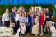 A group of sixteen adults smile and pose together outdoors at an event during the evening, surrounded by plants and palm trees, with tables and white chairs in the foreground—capturing the moment with expert event photography.
