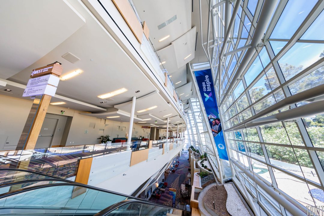 A modern, spacious convention center interior with high ceilings, large windows, natural light, and banners reading "Connect. Inspire." A Corporate Photographer captures people seated by the windows as an escalator leads to the lower level.