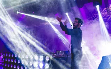 A DJ wearing headphones performs on stage, gesturing with one hand, surrounded by colorful lights and smoke effects, creating a vibrant party atmosphere.