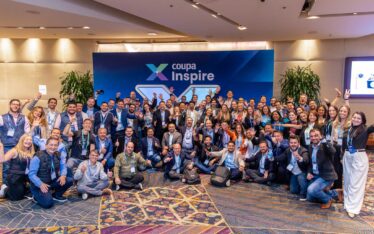 A large, diverse group of professionally dressed people smile and pose together at an indoor event in front of a blue "Coupa Inspire" banner. Many display peace signs and wear conference badges.