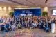 A large, diverse group of professionally dressed people smile and pose together at an indoor event in front of a blue "Coupa Inspire" banner. Many display peace signs and wear conference badges.